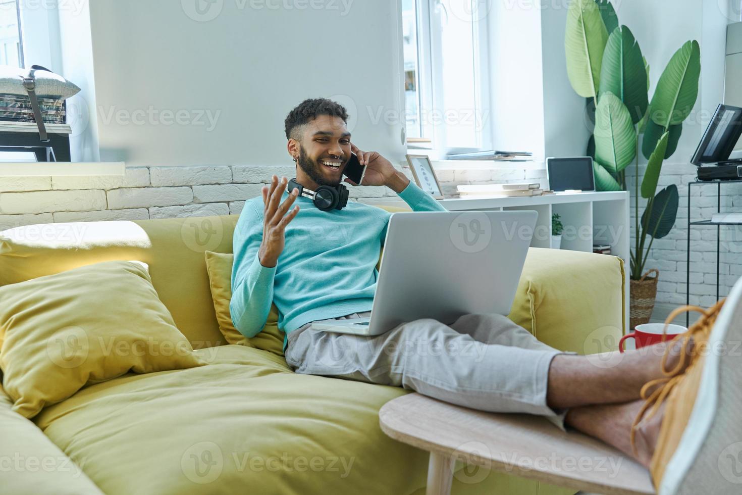 Cheerful African man using laptop and talking on mobile phone while sitting on the couch photo