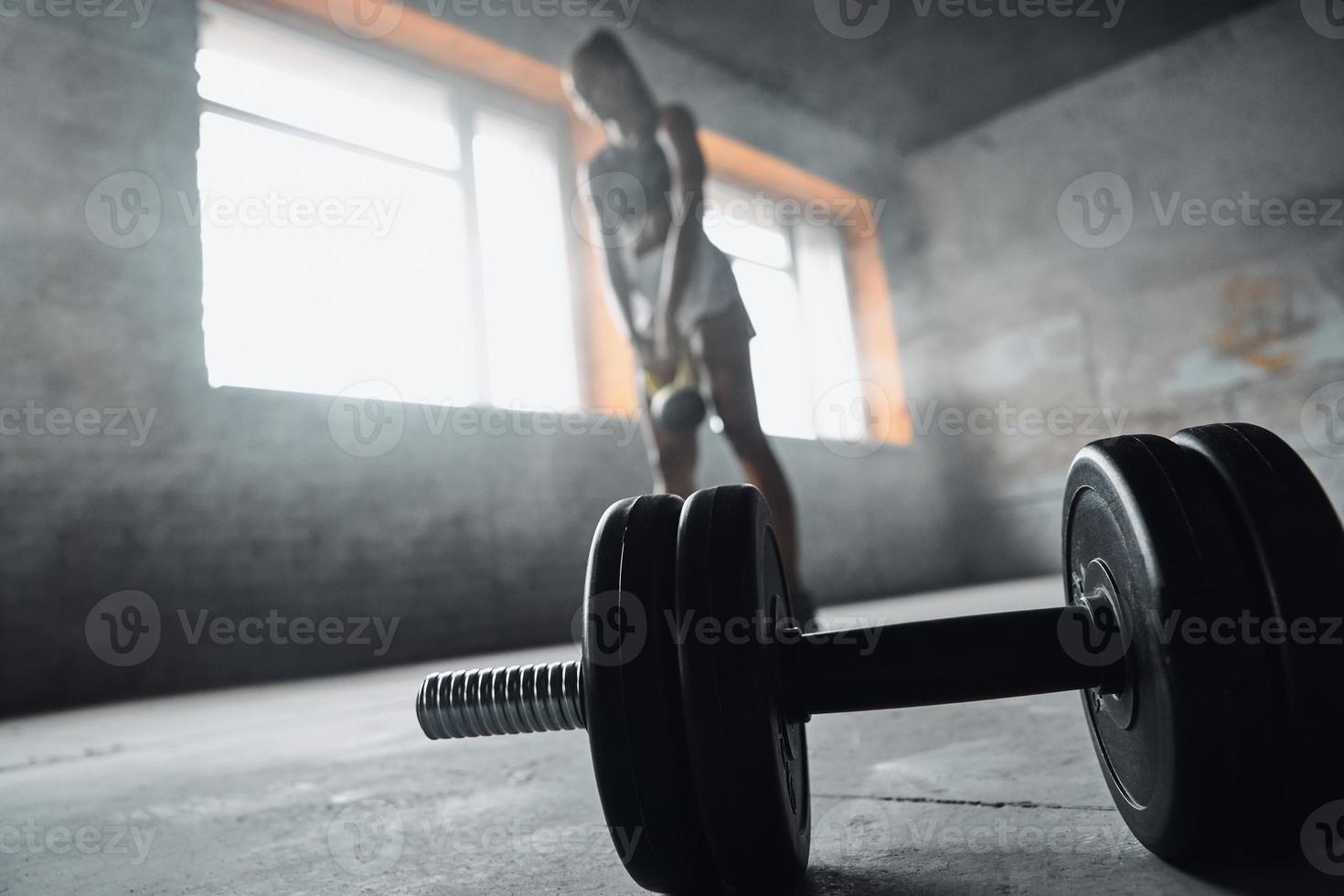 Concentrated young African woman exercising with kettlebell while dumbbell laying on foreground photo