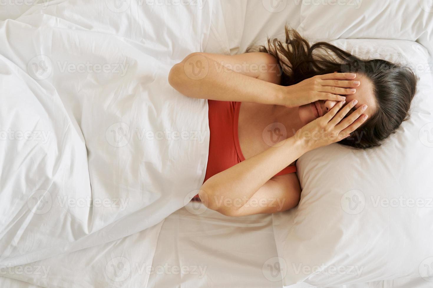 Top view of depressed young woman covering face with hands while lying in bed photo