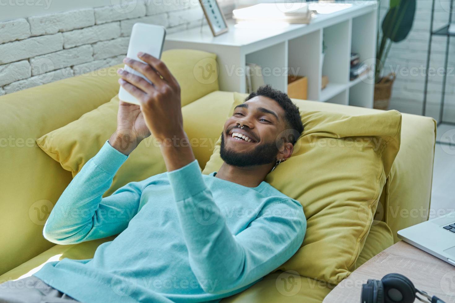 Happy African man using smart phone while relaxing on the couch at home photo