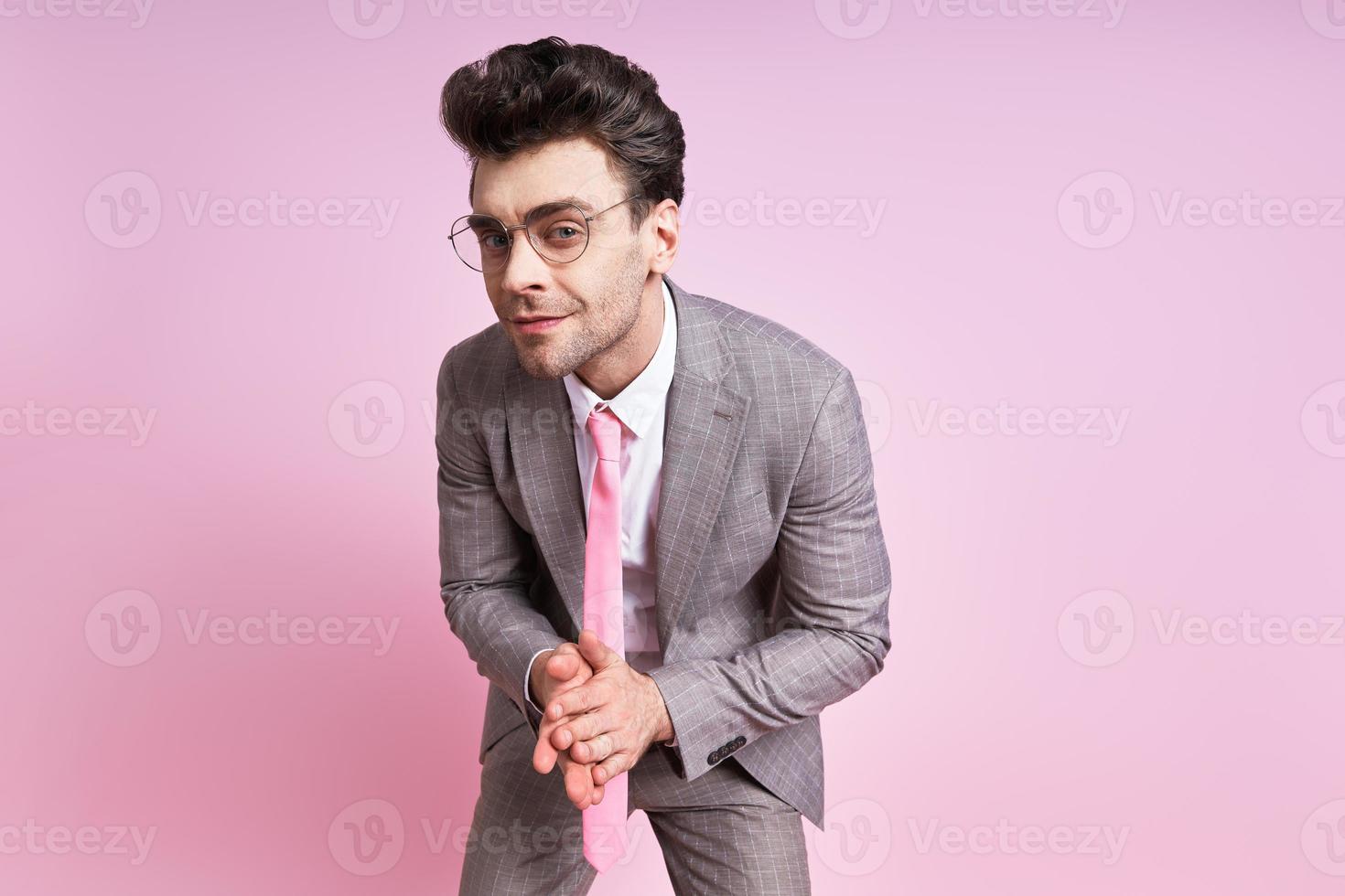 Confident man in full suit keeping hands clasped while standing against pink background photo