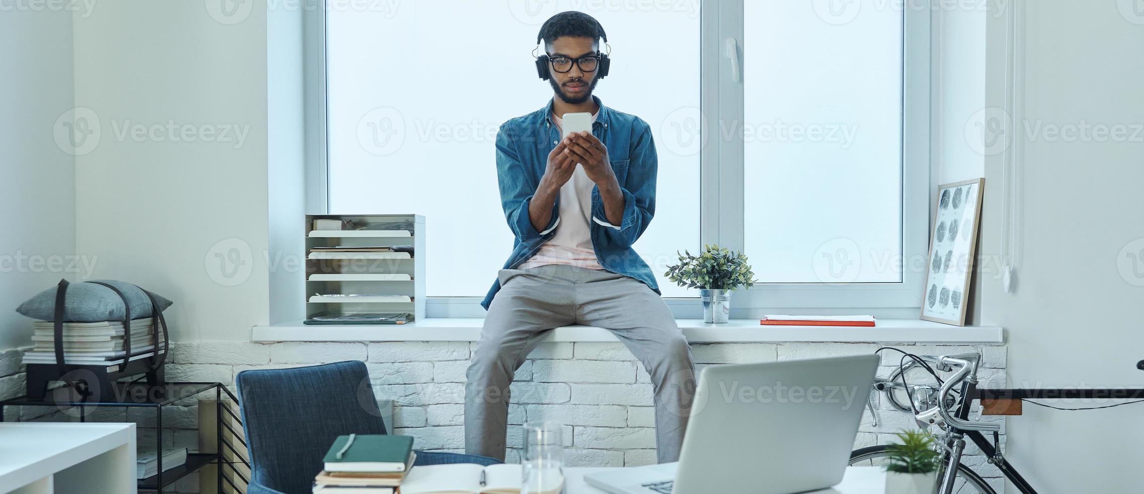 Confident African man using smart phone while sitting on the window sill in office photo