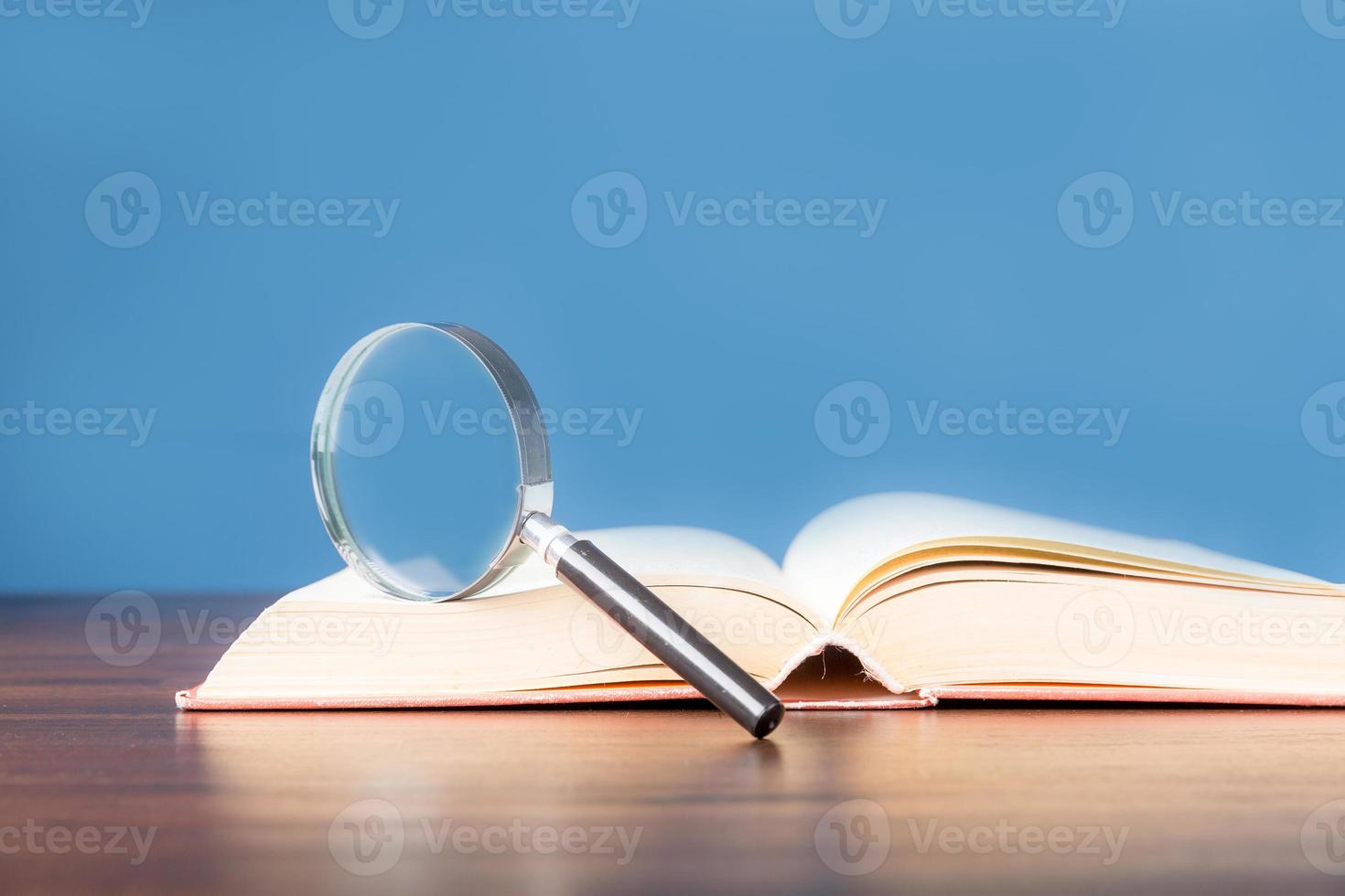 open book with magnifying glass on wooden desk in information library of school or university, concept for education,reading , study, copy space and blue background. photo