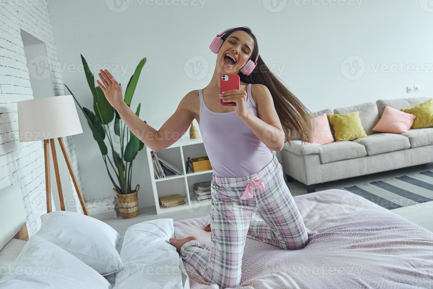 Young woman using smart phone like microphone while dancing on bed at home photo