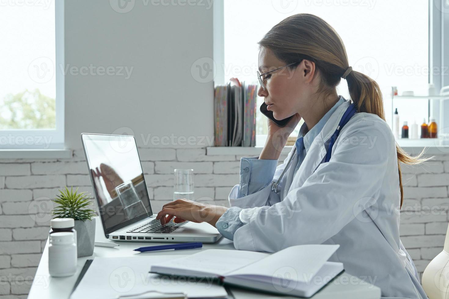 Confident female doctor talking on phone and using computer while sitting at the medical office photo