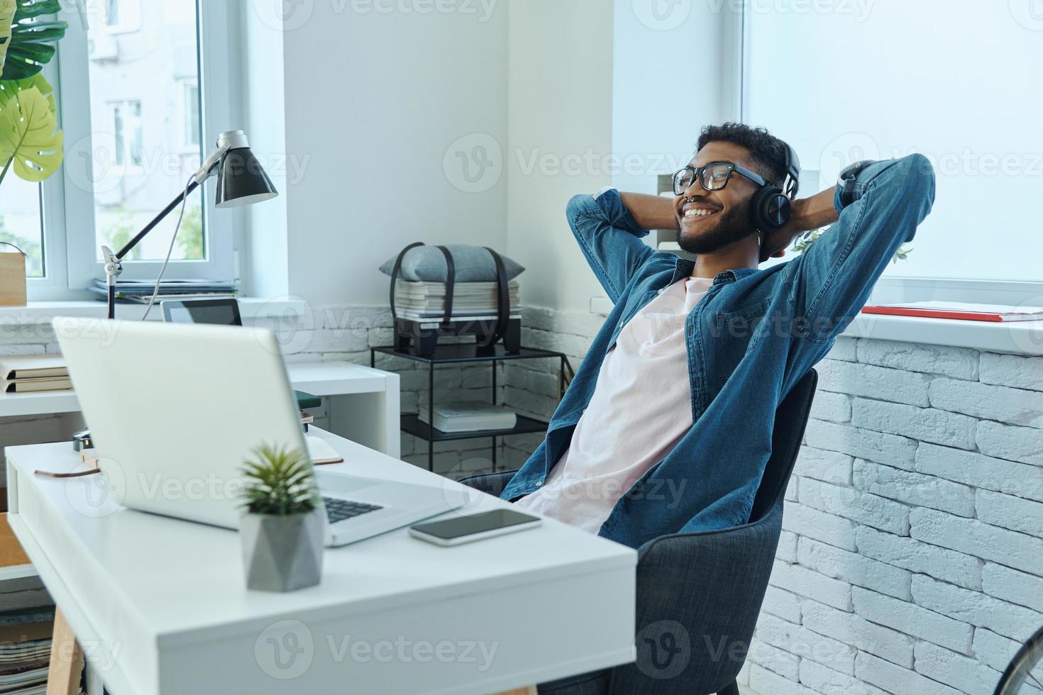 Relaxed African man in headphones holding hands behind head while sitting at working place photo