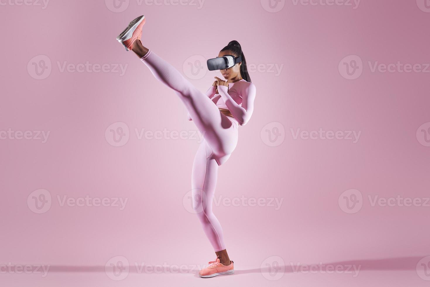 Woman in virtual reality headset practicing in kicking while standing against pink background photo