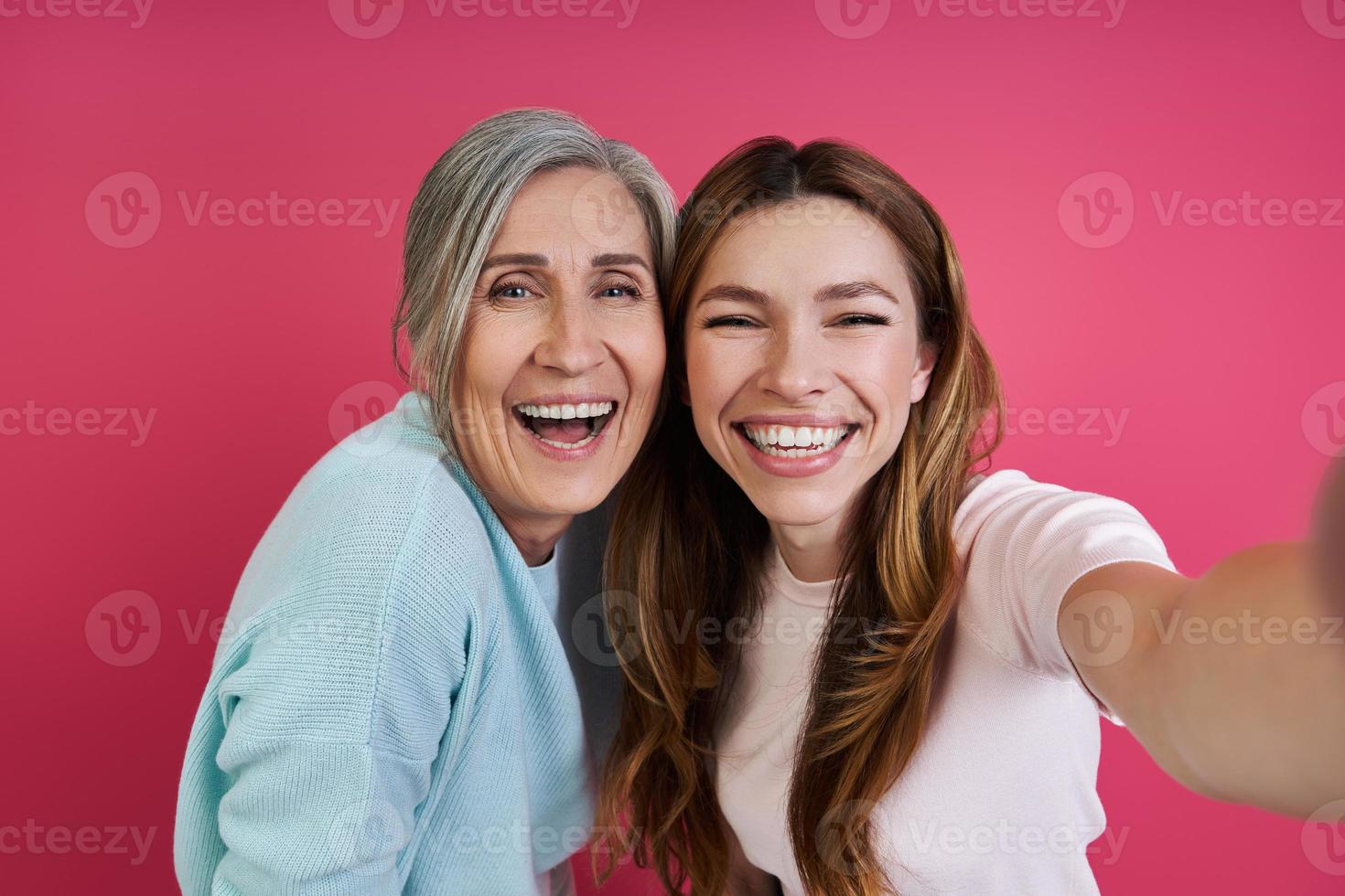 feliz madre mayor y su hija adulta haciendo selfie contra fondo rosa foto