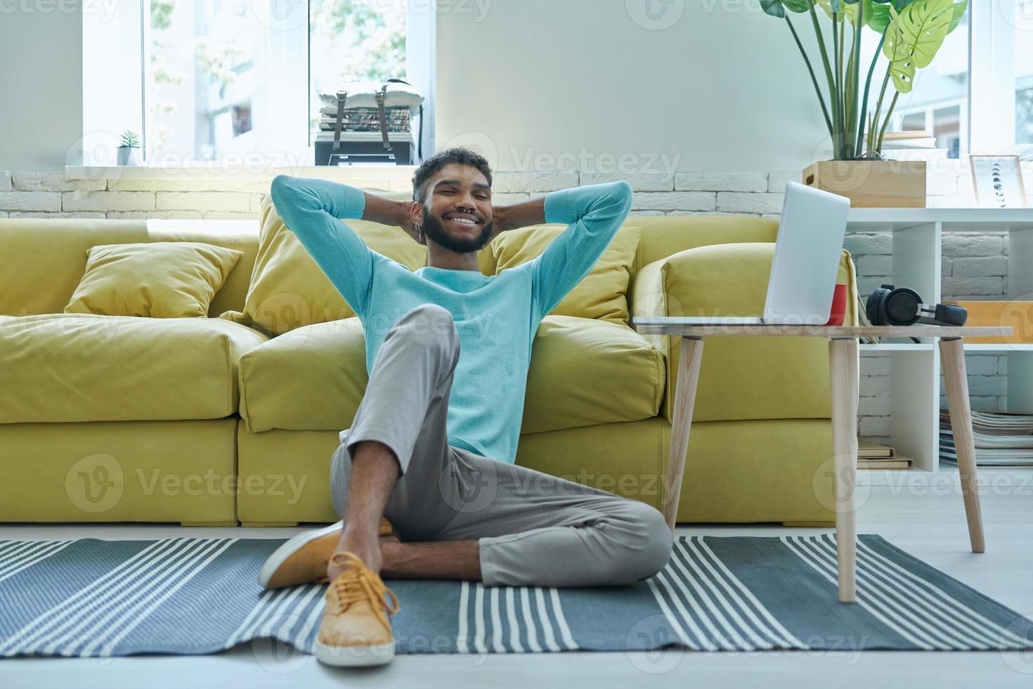 Relaxed African man holding hands behind head and smiling while sitting on the floor at home photo