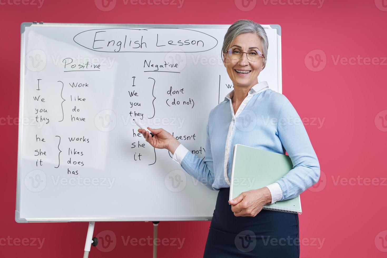 Cheerful senior woman teaching English language near the whiteboard against pink background photo