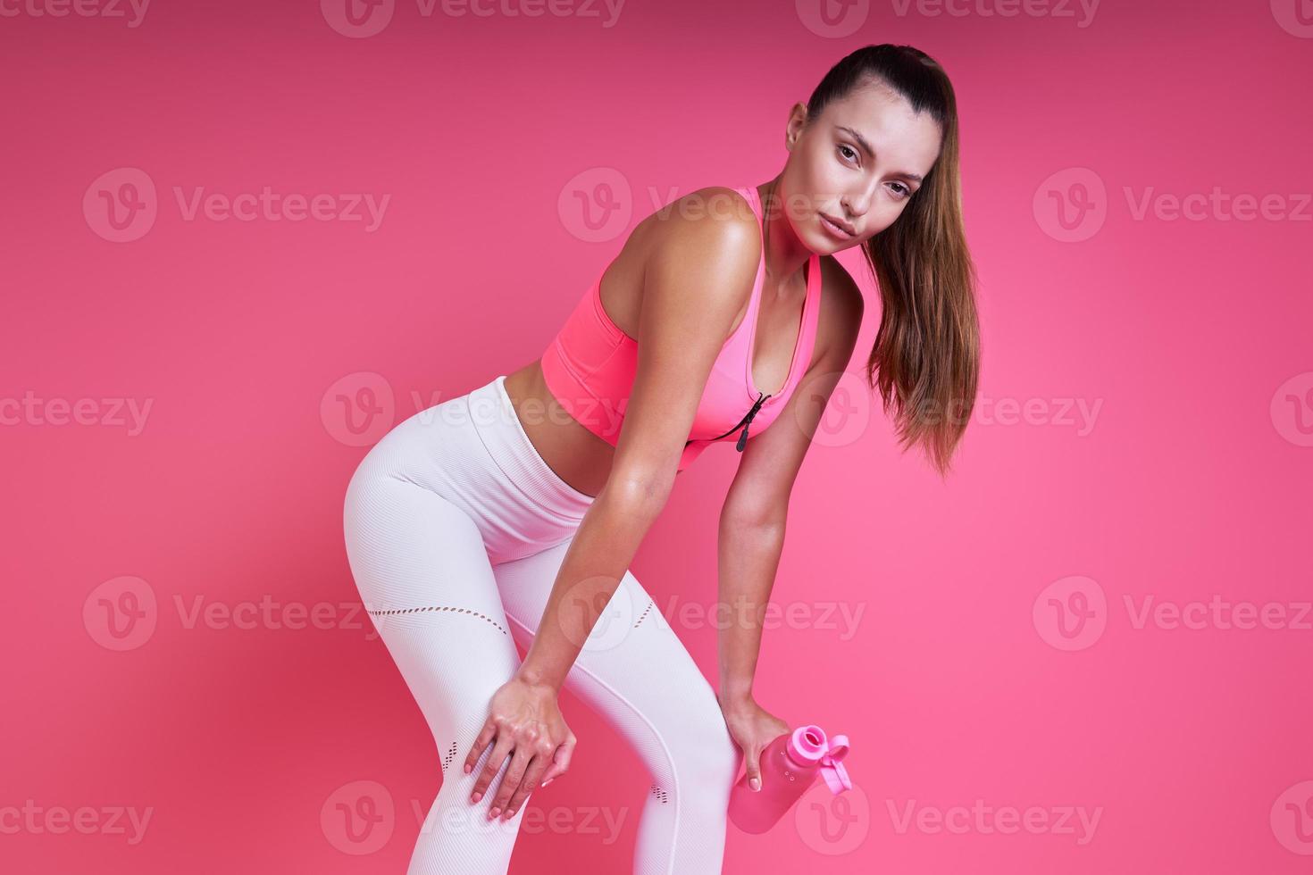 Relaxed young woman leaning on knees while standing against pink background photo