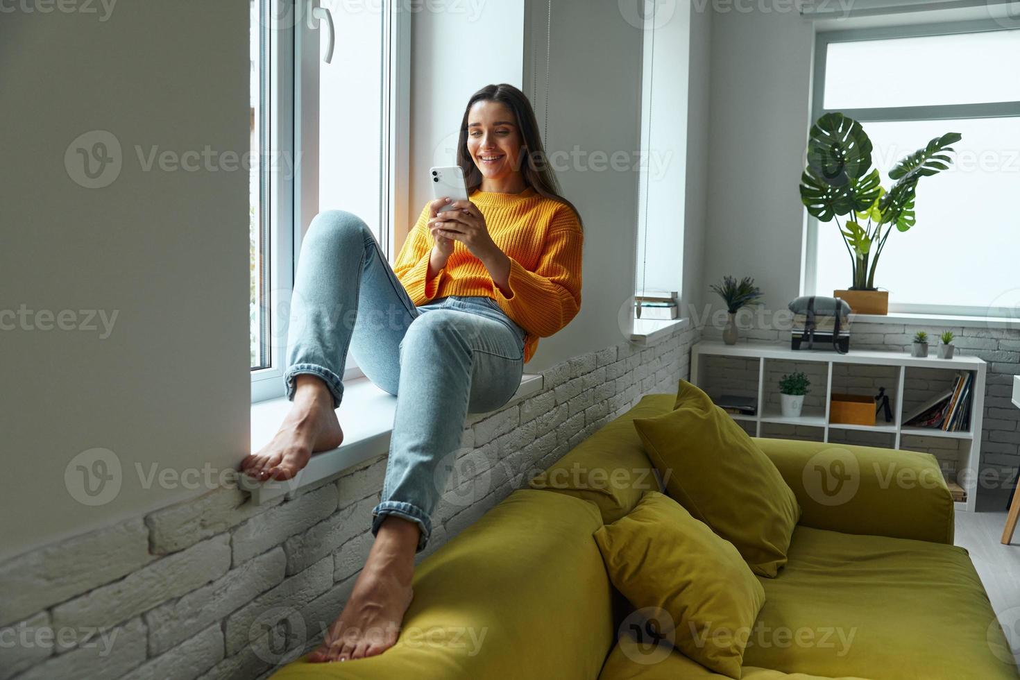 Happy young woman using smart phone while sitting on the window sill at home photo