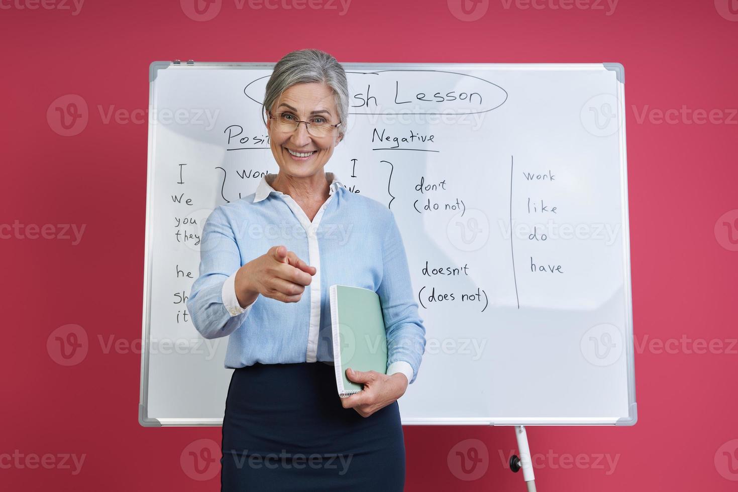 Cheerful female teacher pointing camera while standing near the whiteboard against pink background photo