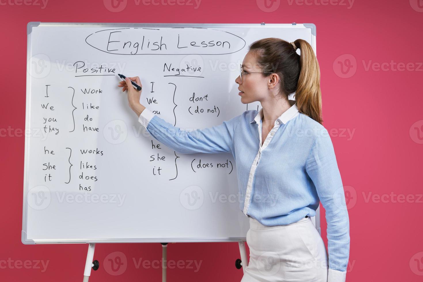 Young woman teaching English language while standing near the whiteboard against pink background photo