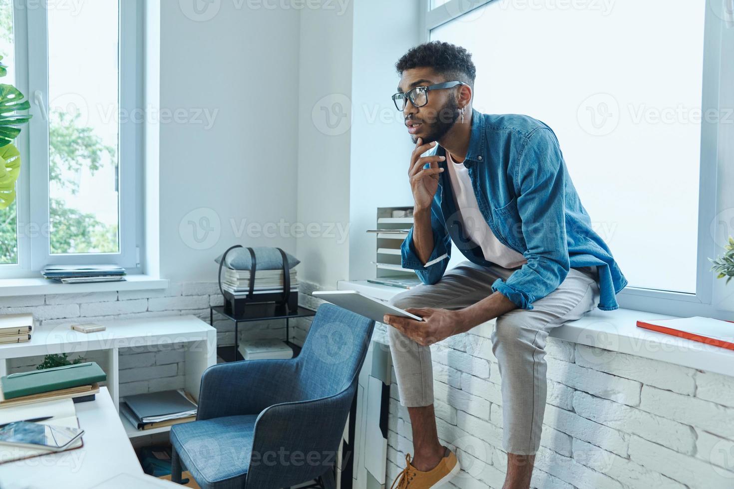 Thoughtful young man holding digital tablet while sitting on the window sill photo
