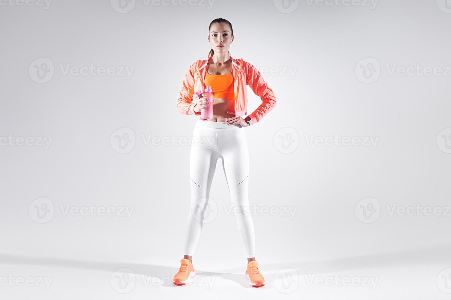 Full length of beautiful young woman in sports clothing drinking water against white background photo