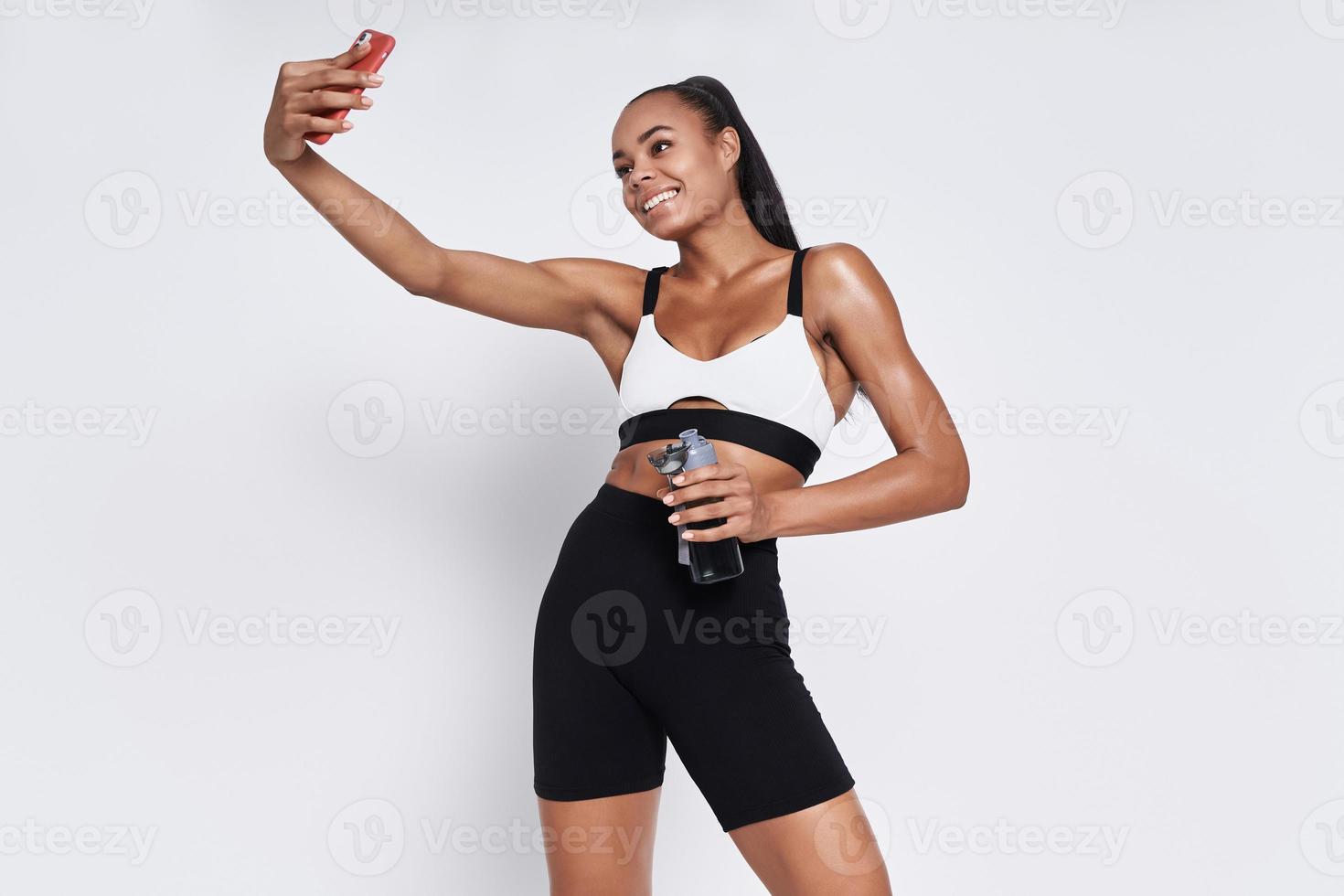 Happy African woman in sports clothing holding bottle and making selfie against white background photo