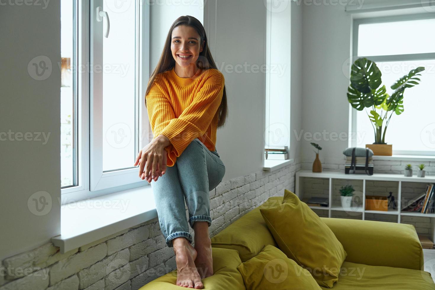 mujer joven feliz mirando a la cámara mientras se sienta en el alféizar de la ventana en casa foto