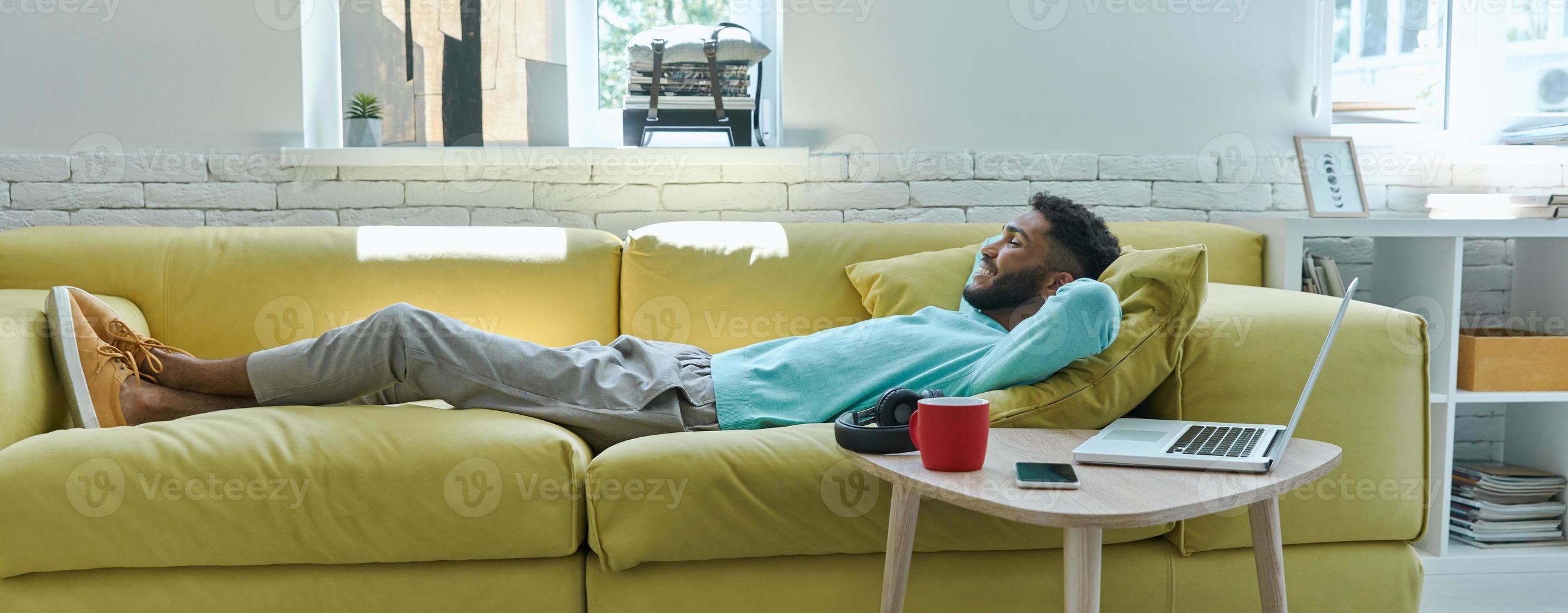 Carefree African man holding hand behind head while relaxing on the couch at home photo