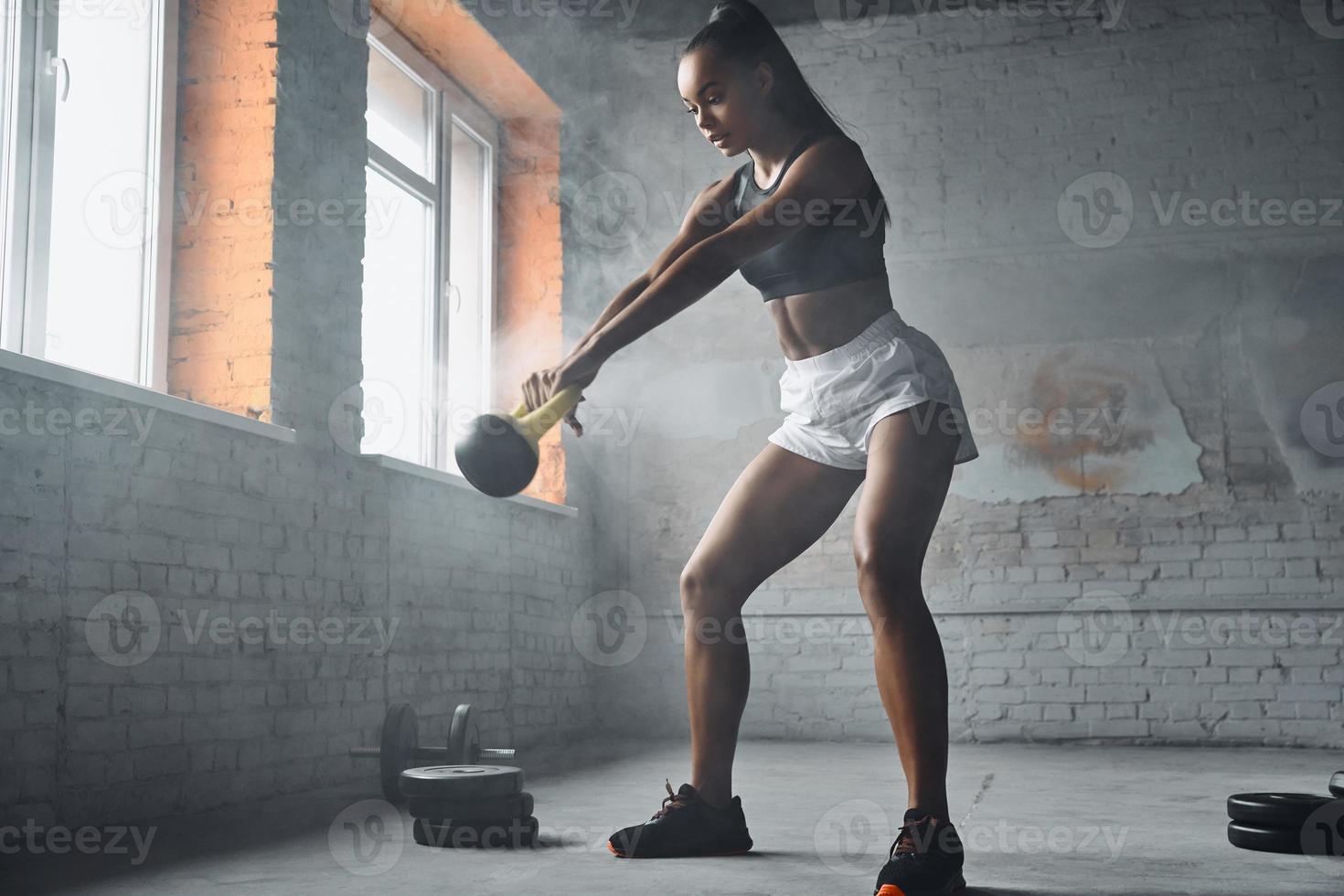 Full length of beautiful young African woman exercising with kettlebell in gym photo