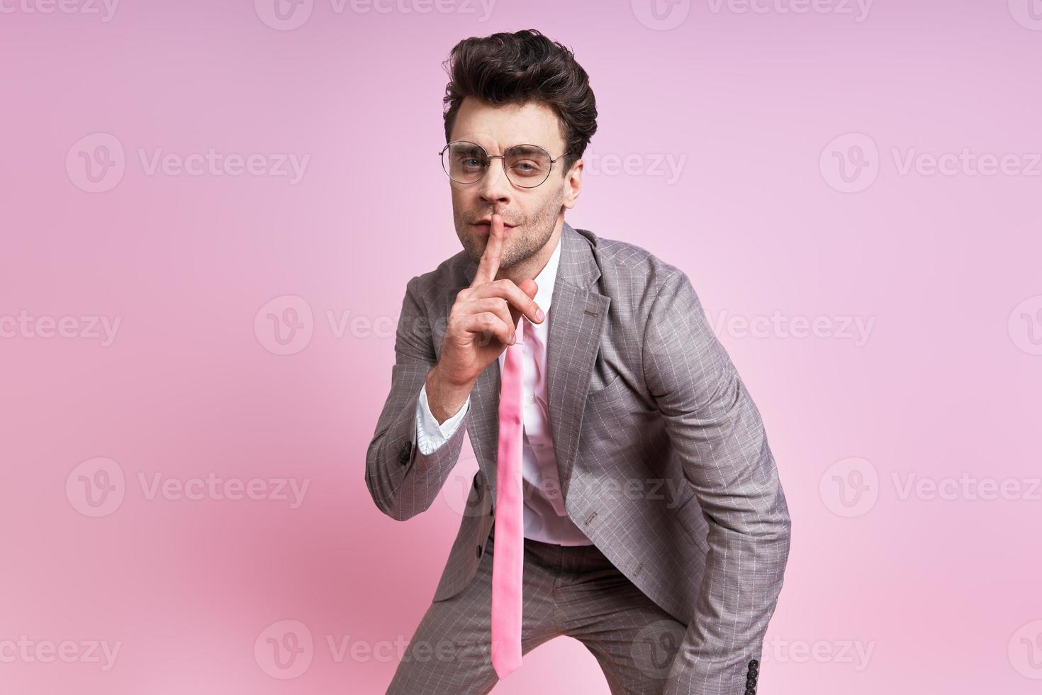 Confident man in full suit holding finger on lips while standing against pink background photo