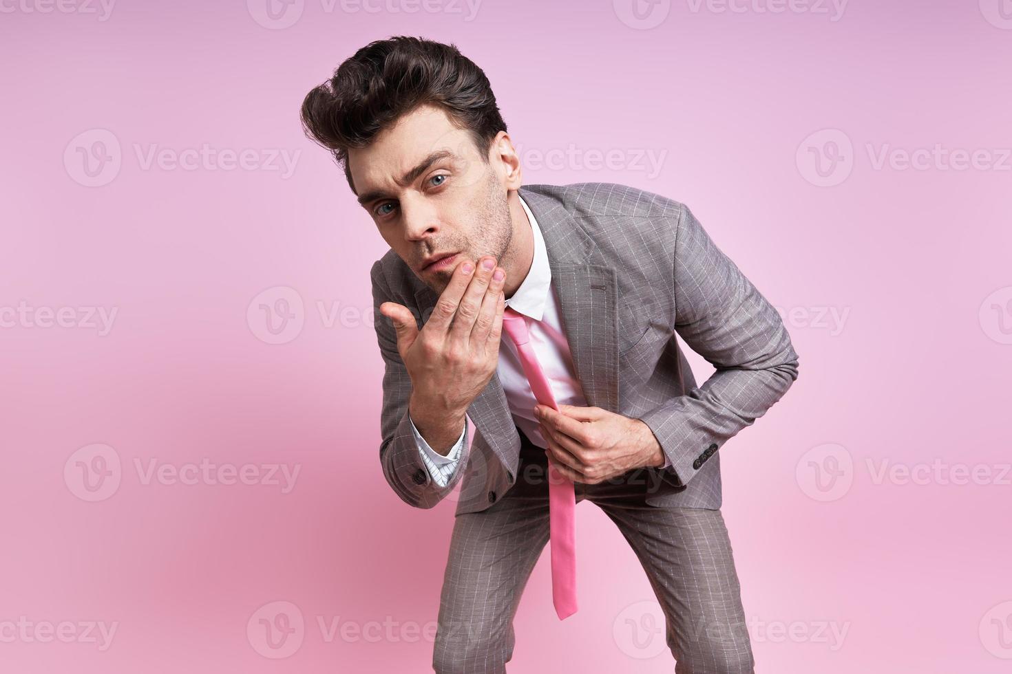 Handsome man in full suit touching his face while standing against pink background photo
