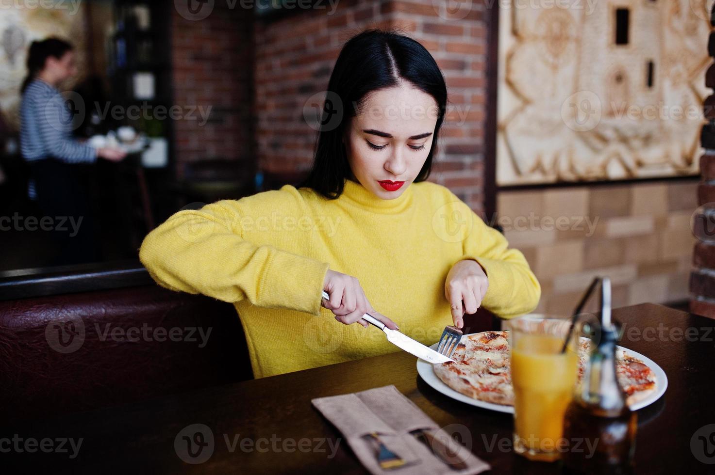 Funny brunette girl in yellow sweater eating pizza at restaurant. photo