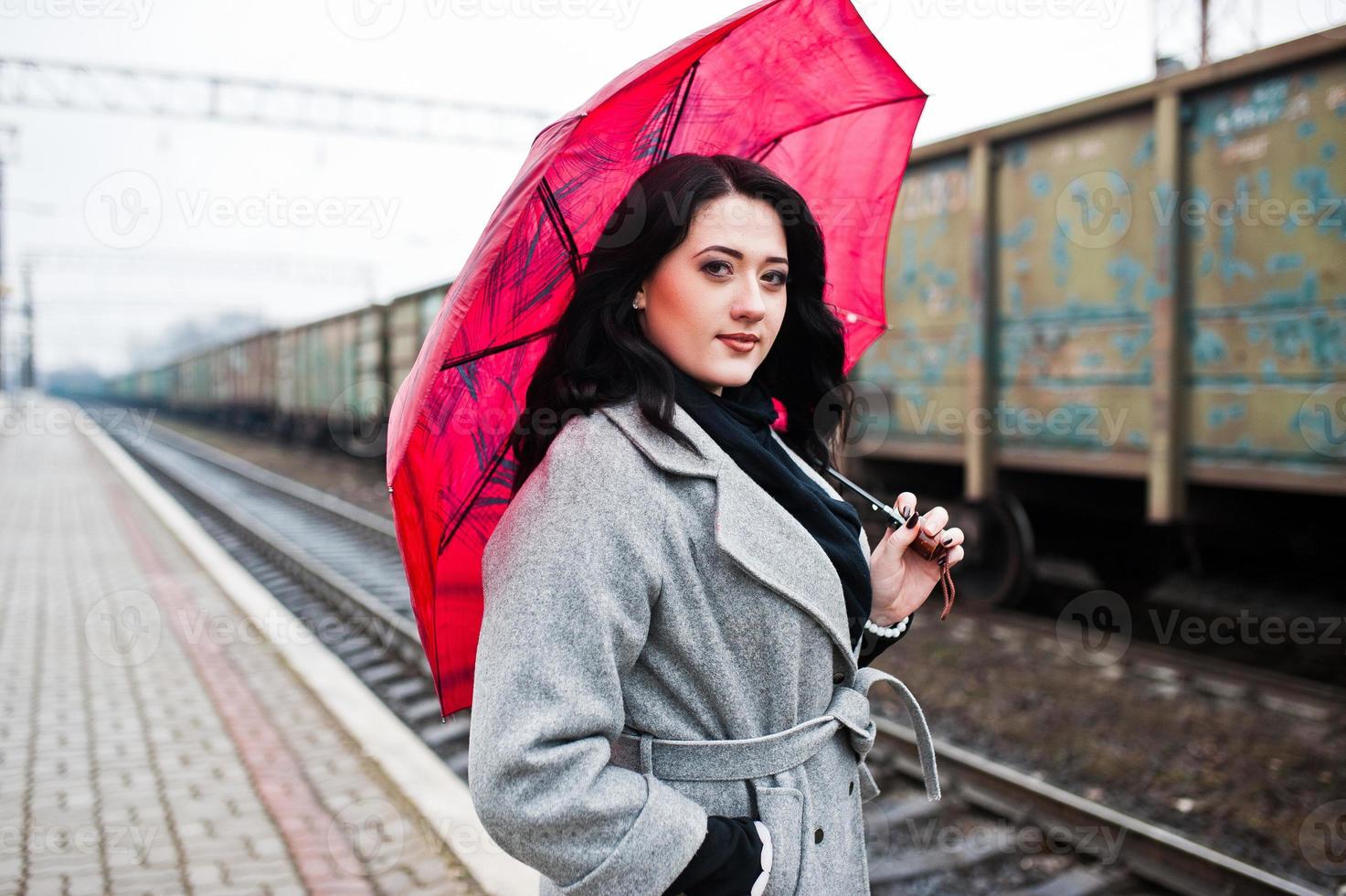 Brunette girl in gray coat with red umbrella in railway station. photo