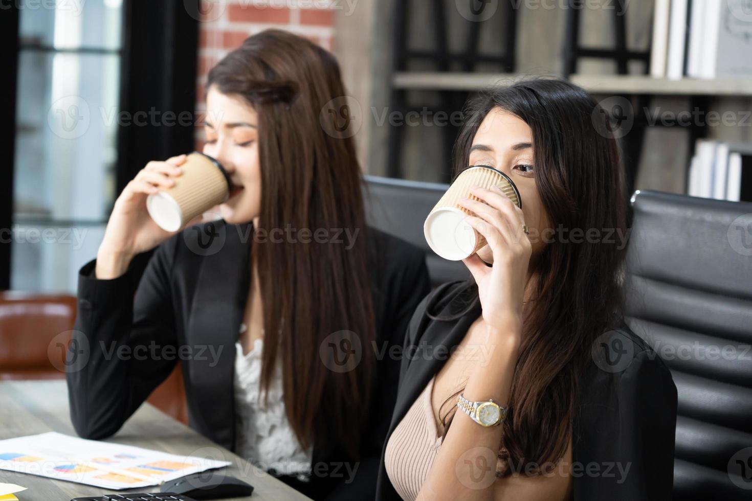 Office coffee break with two female colleagues sitting chatting over cups of coffee. Asian business woman holding coffee cup in office. photo