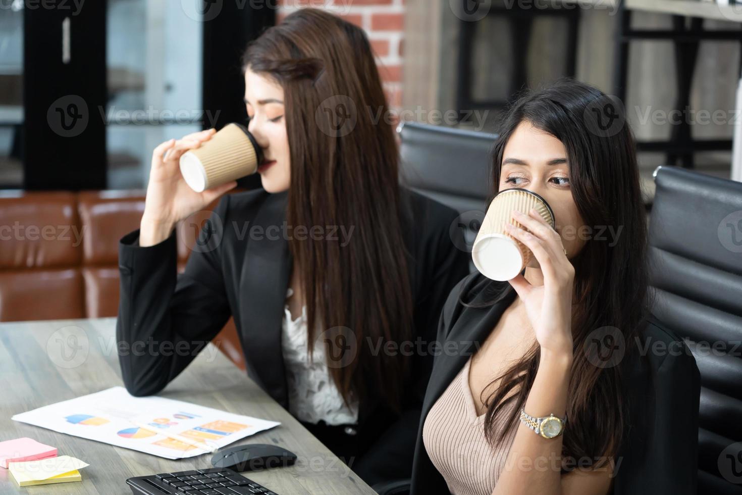 Office coffee break with two female colleagues sitting chatting over cups of coffee. Asian business woman holding coffee cup in office. photo