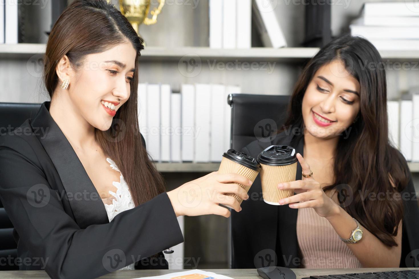 Office coffee break with two female colleagues sitting chatting over cups of coffee. Asian business woman holding coffee cup in office. photo