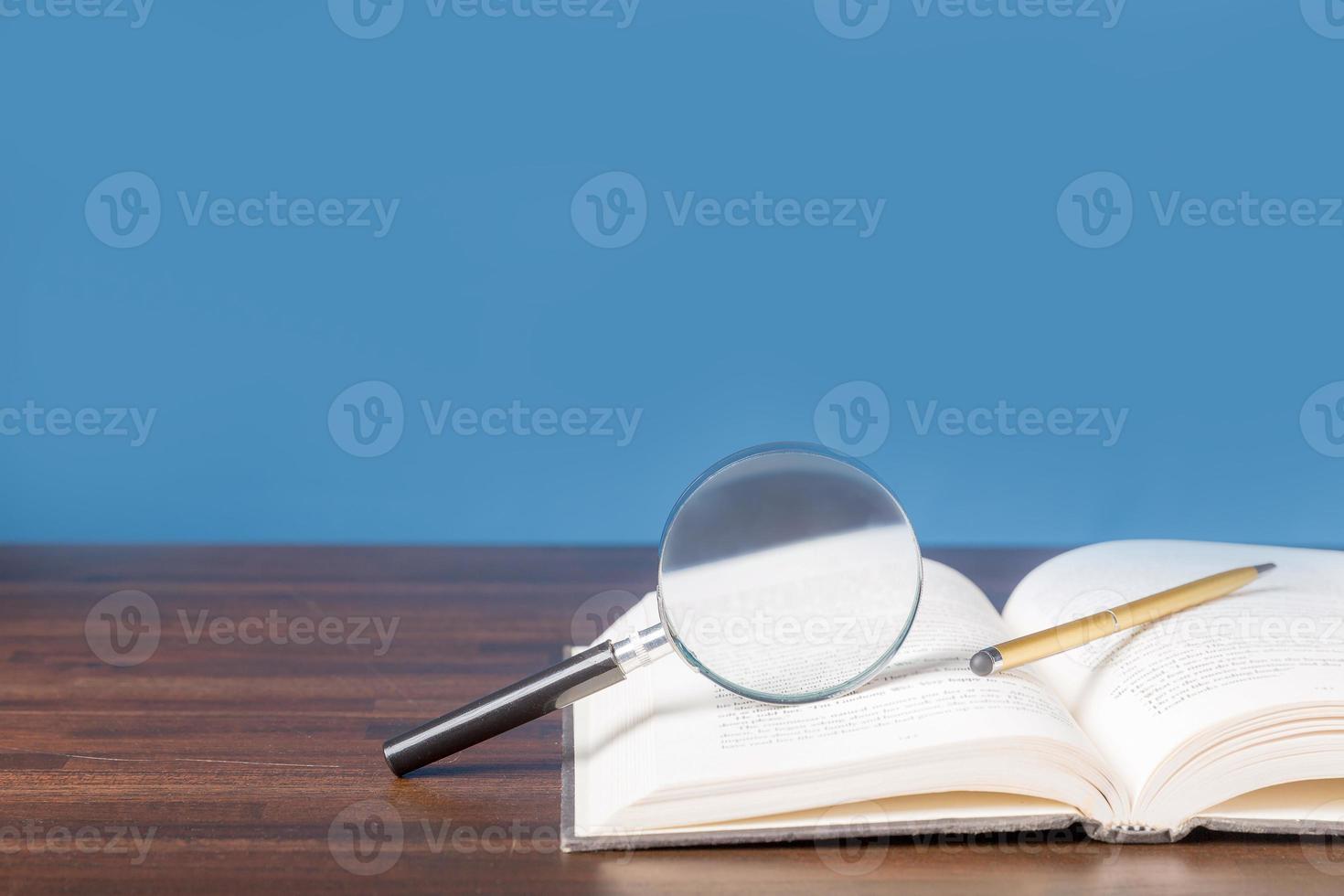 open book with magnifying glass on wooden desk in information library of school or university, concept for education,reading , study, copy space and blue background. photo