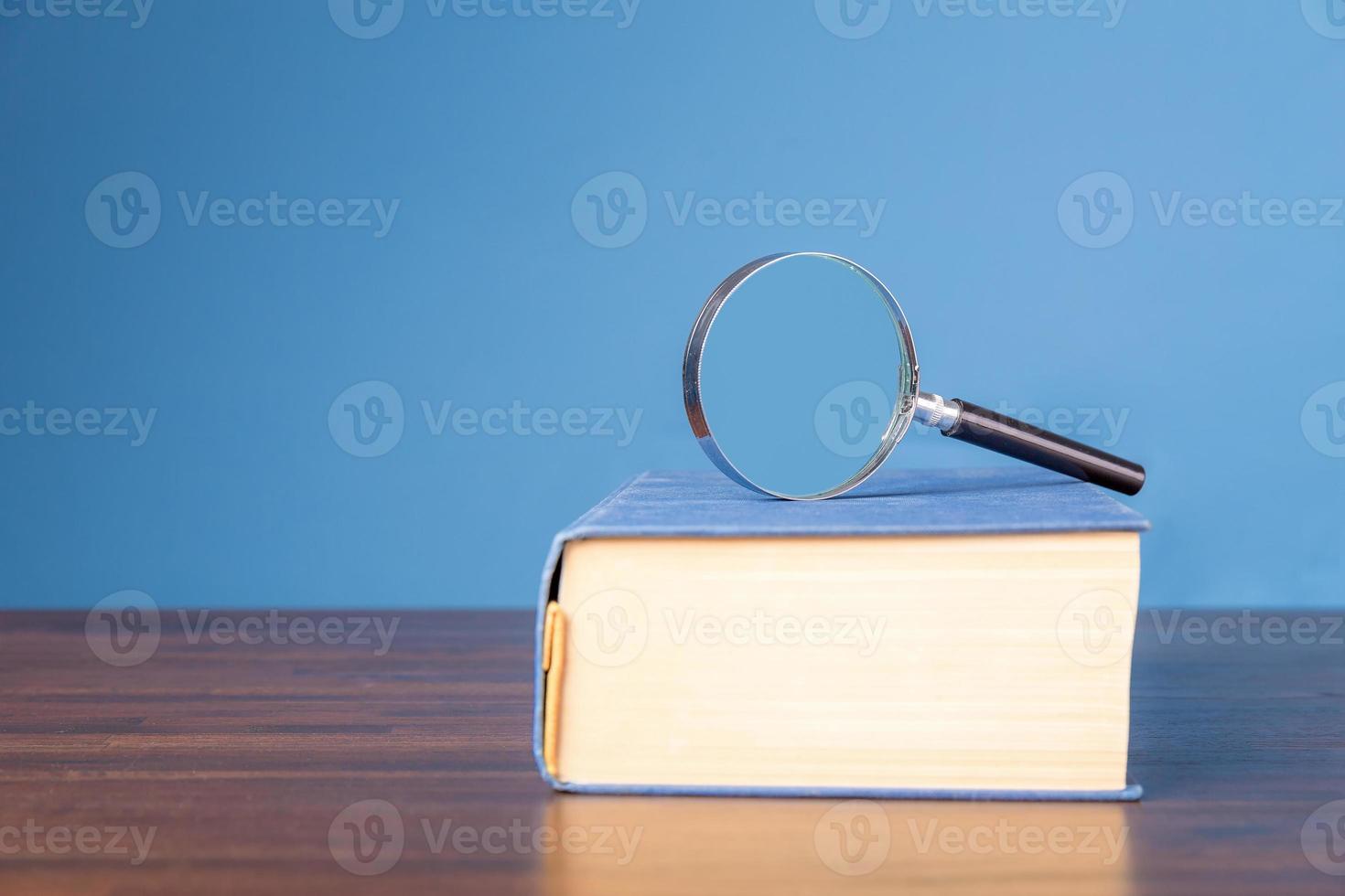 Old book with magnifying glass on wooden desk in information library of school or university, concept for education, reading , study, copy space and blue background. photo