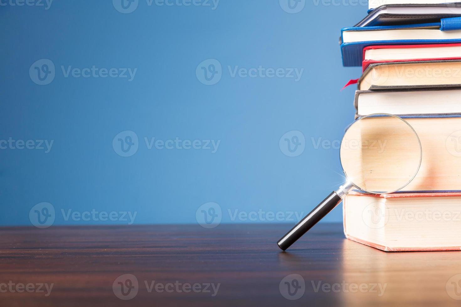 Stack book with magnifying glass on wooden desk in information library of school or university, concept for education, reading, study, copy space on blue background. photo