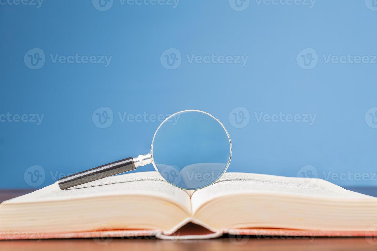 open book with magnifying glass on wooden desk in information library of school or university, concept for education,reading , study, copy space and blue background. photo