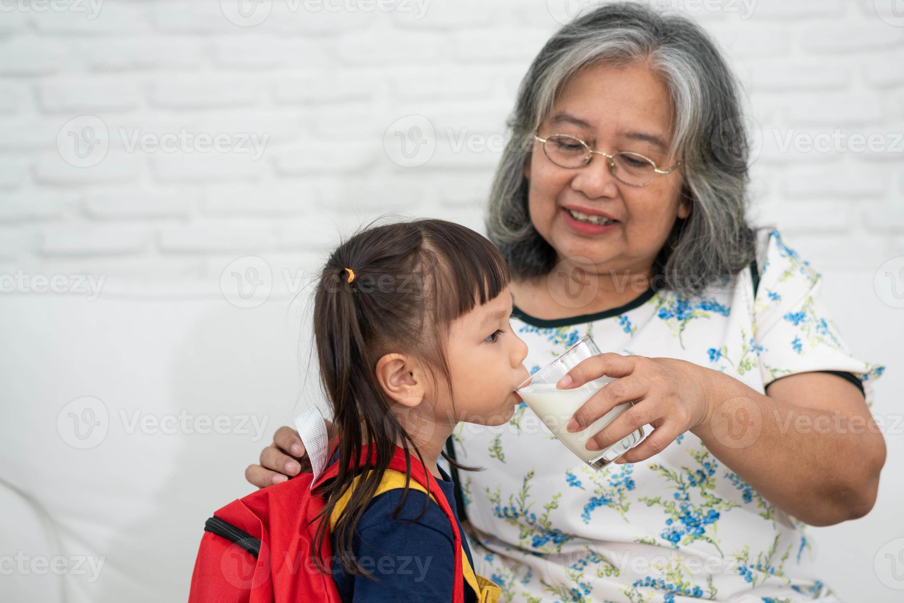 Happy Elderly Asian Grandma Sits Beside Her Granddaughter And Feeds Fresh Milk From Glass For