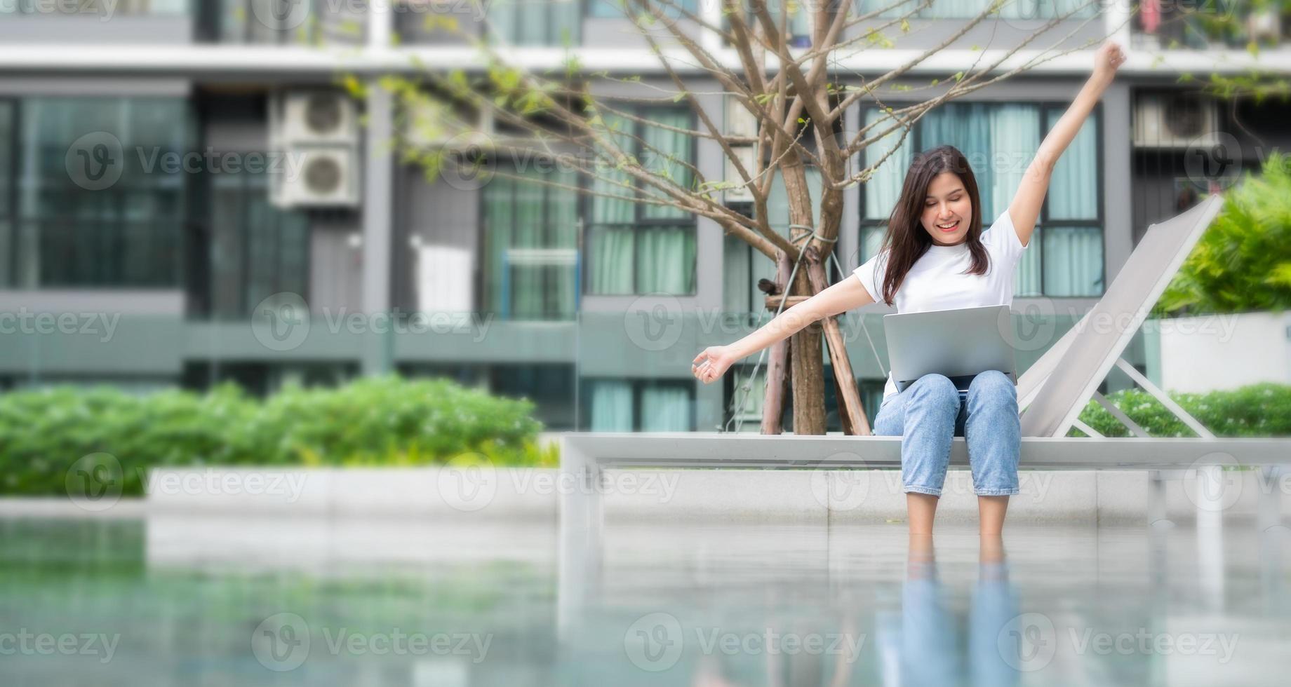 Happy young entrepreneur woman sitting on tanning bed beside pool and using laptop computer for remote online working digital, online business project in quiet yard of resort house, Work on vacation photo