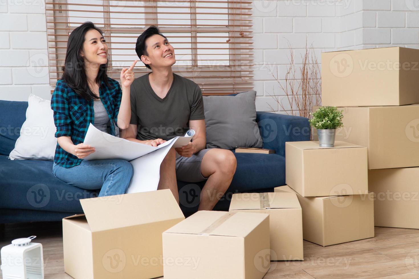 Smiling young Asian happy couple hold blueprint for home decoration ideas at moving day in their new home after buying real estate. Concept of starting a new life for a newly married couple. photo