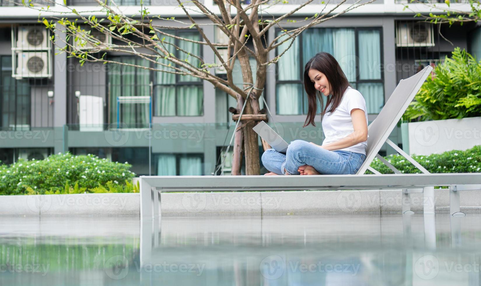 Happy young entrepreneur woman sitting on tanning bed beside pool and using laptop computer for remote online working digital, online business project in quiet yard of resort house, Work on vacation photo