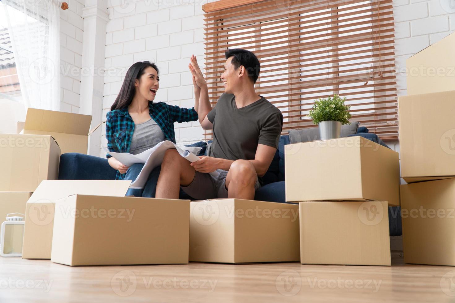 Smiling young Asian happy couple hold blueprint for home decoration ideas at moving day in their new home after buying real estate. Concept of starting a new life for a newly married couple. photo
