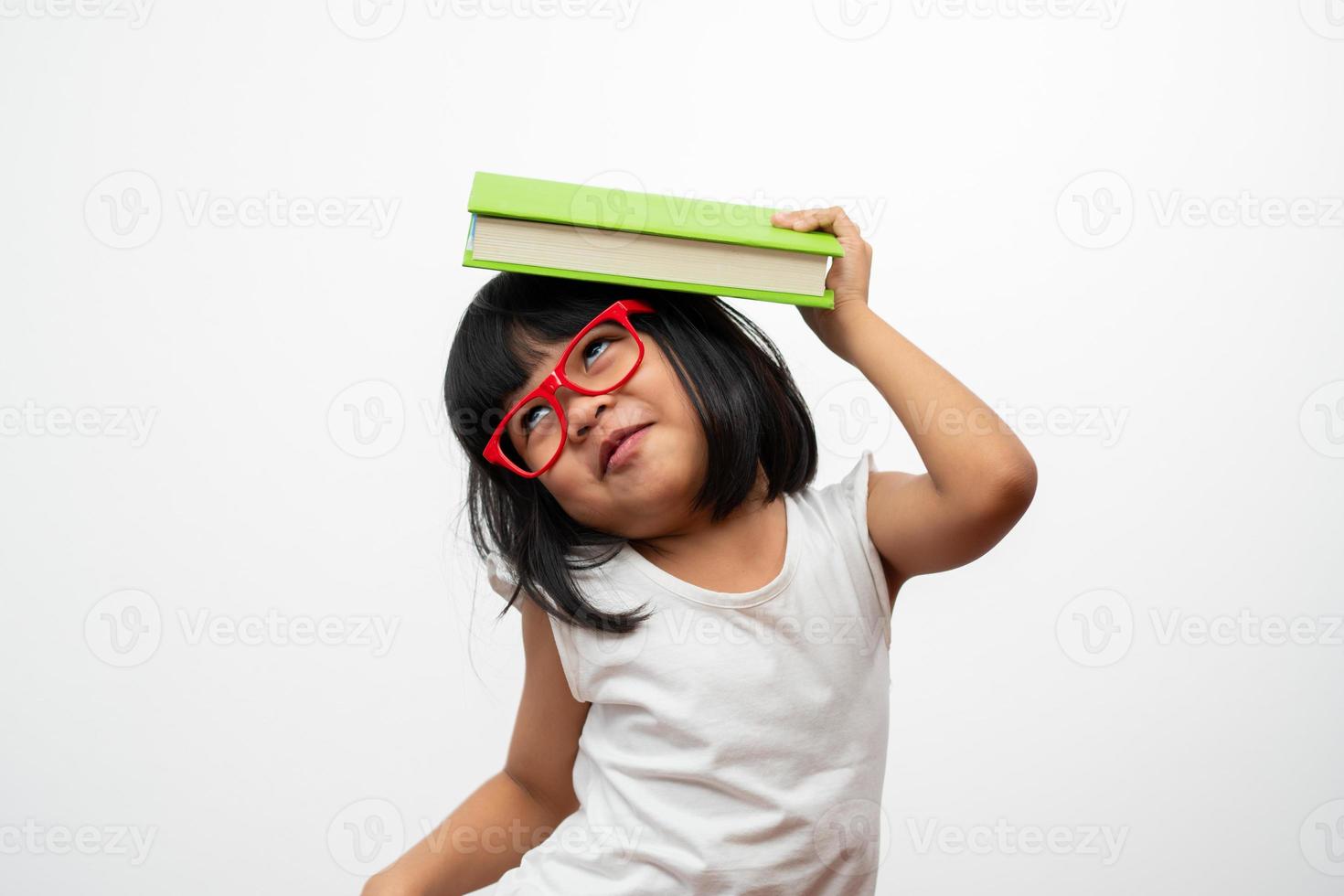 una niña preescolar asiática divertida y feliz con gafas rojas sosteniendo un libro verde en la cabeza, con un fondo blanco aislado. concepto de niño escolar y educación en primaria y preescolar foto