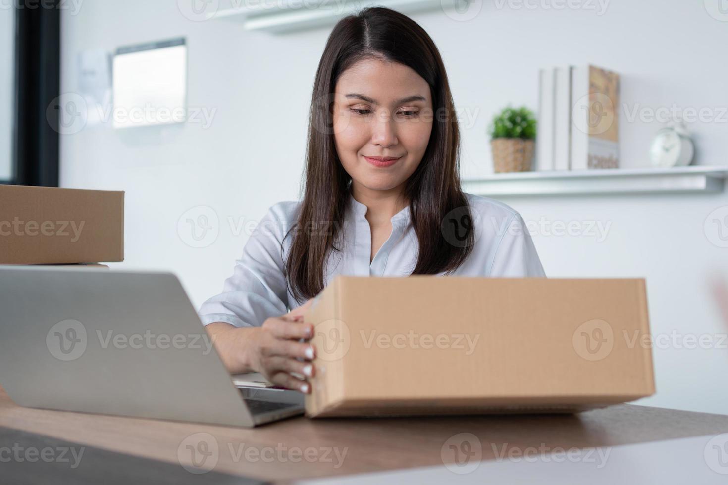 Happy young Asian woman entrepreneur, Hold package and Smile for sale success of online shopping store at home office, Concept of merchant, small business, online business and eCommerce. photo