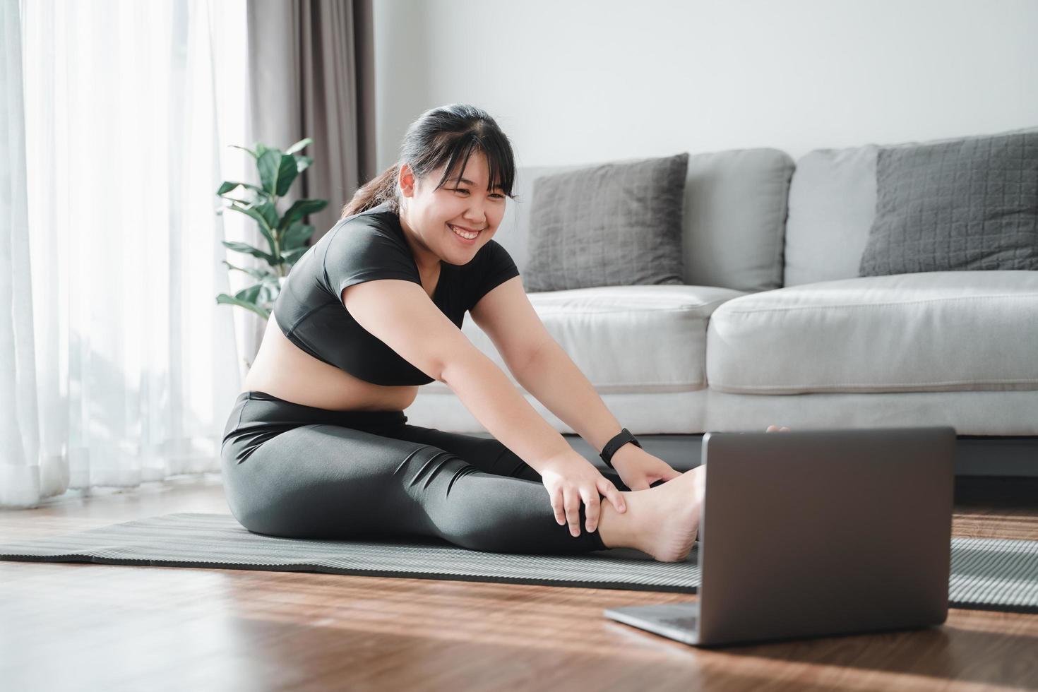 una mujer gordita asiática sentada en el suelo en la sala de estar practica una lección de yoga en línea con la computadora. mujer con clase de entrenamiento de meditación en la computadora portátil. foto