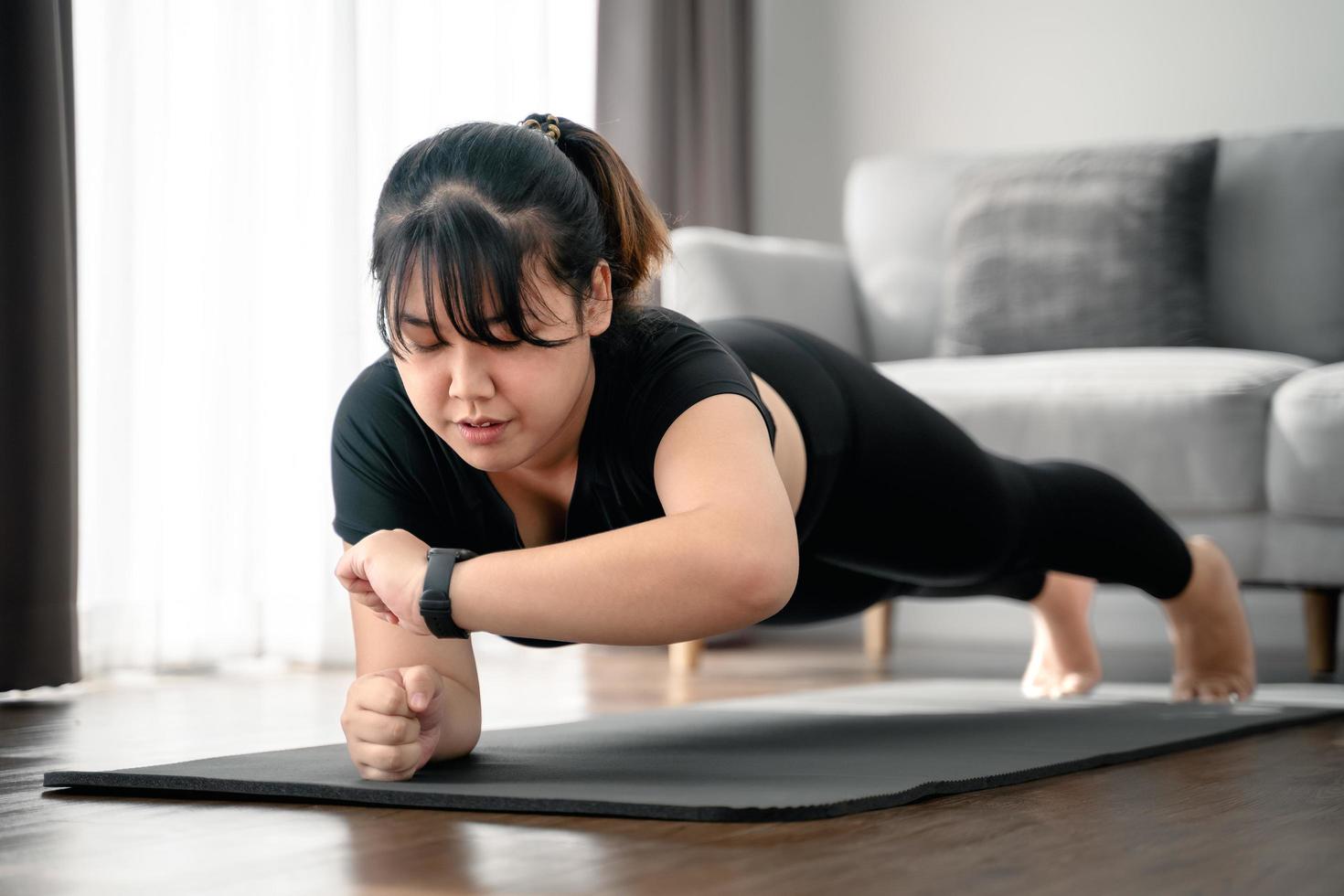 Asian chubby young woman doing planking exercise and looking at smartwatch screen indoors at home . photo