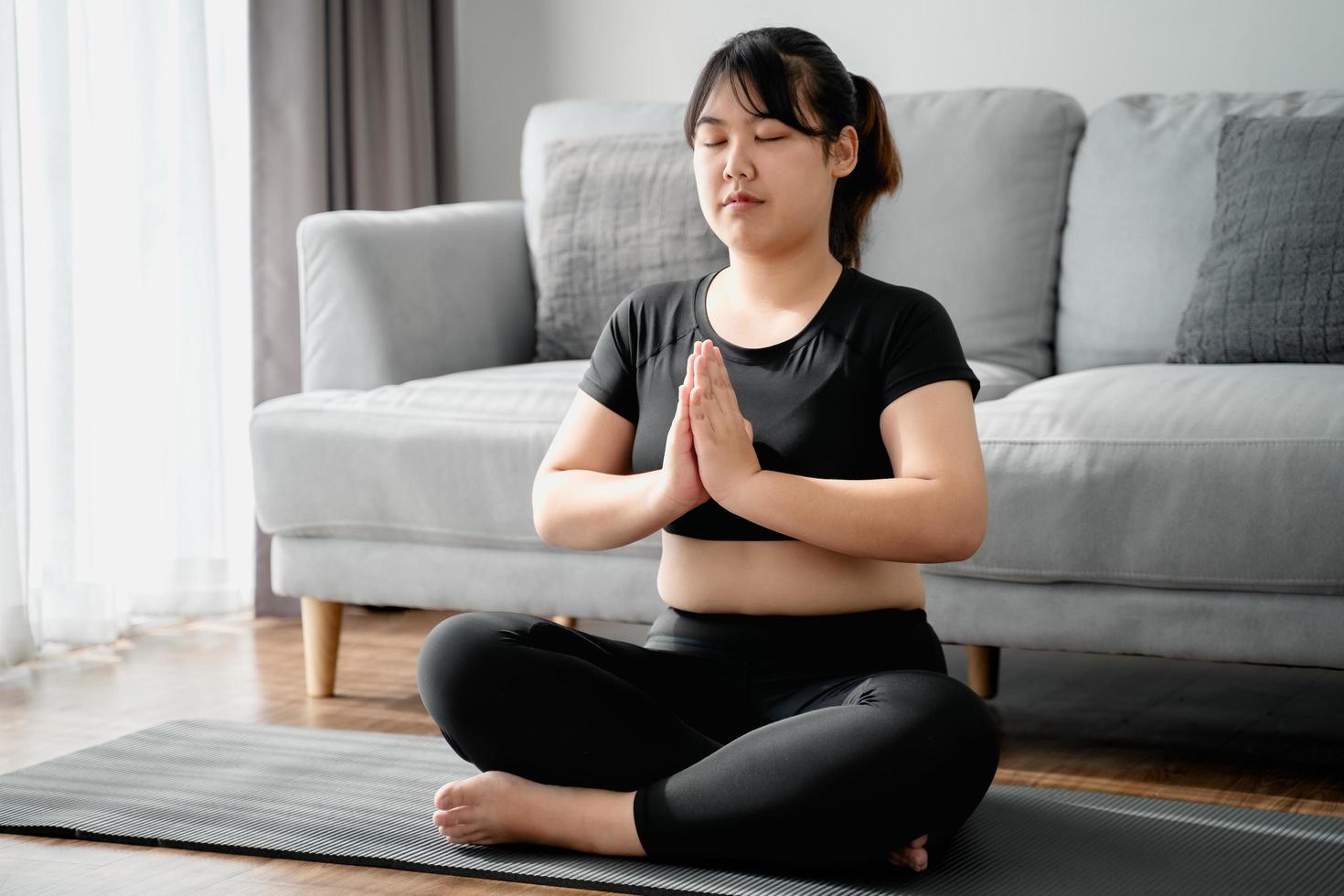 mujer gordita asiática sentada en el suelo en la sala de estar practica la lección de yoga. mujer con clase de entrenamiento de meditación. foto