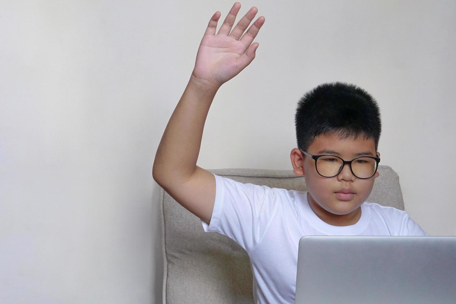 The boy sit on sofa studying through laptop and raises his hand to answer lesson. Education, learning and technology concept. photo