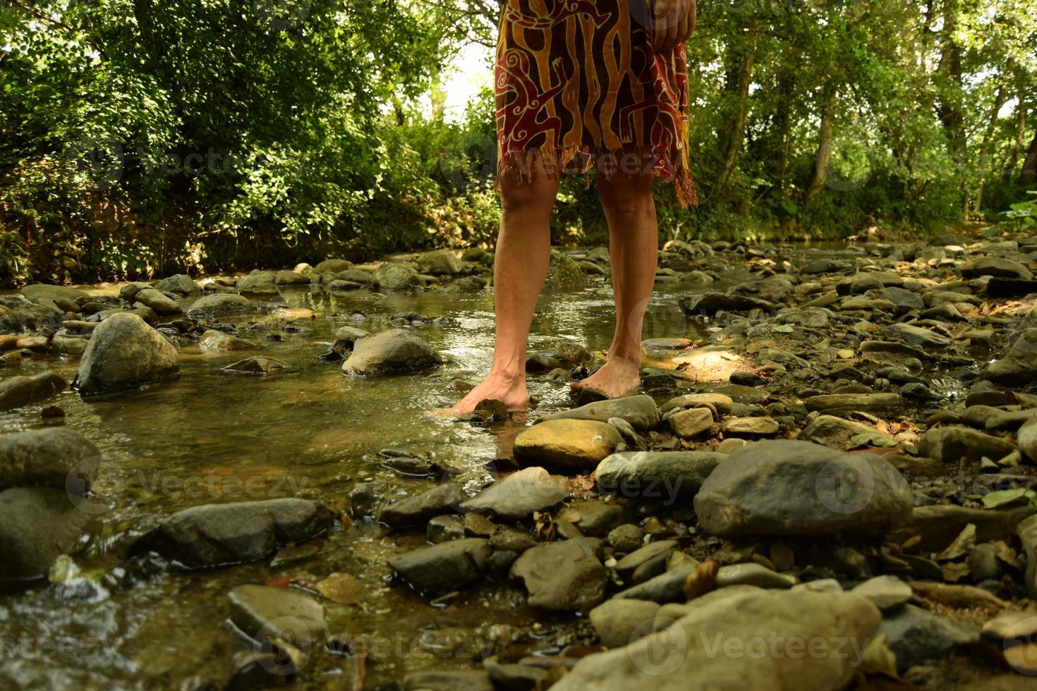 A barefoot woman walking in a clean brook water with rocks and stones around her feet, among greenery, in the shades of trees in a sunny day photo