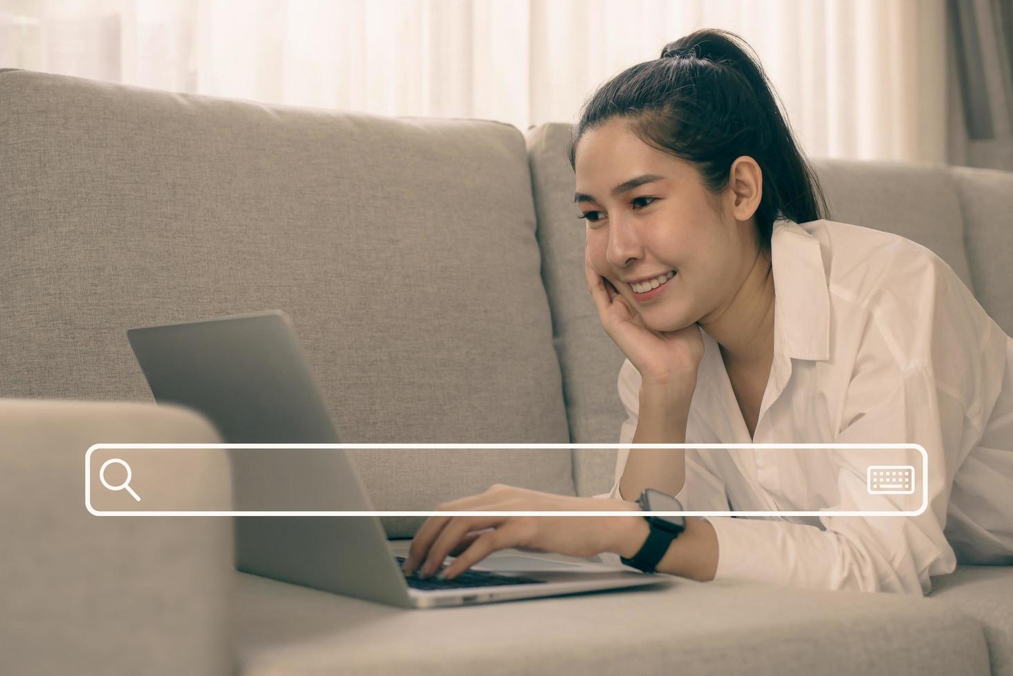 alegre mujer asiática sonriente usando una laptop con un icono de búsqueda mientras se sienta en el sofá en la sala de estar. ella usa su computadora portátil para buscar y disfrutar de las compras en línea. foto