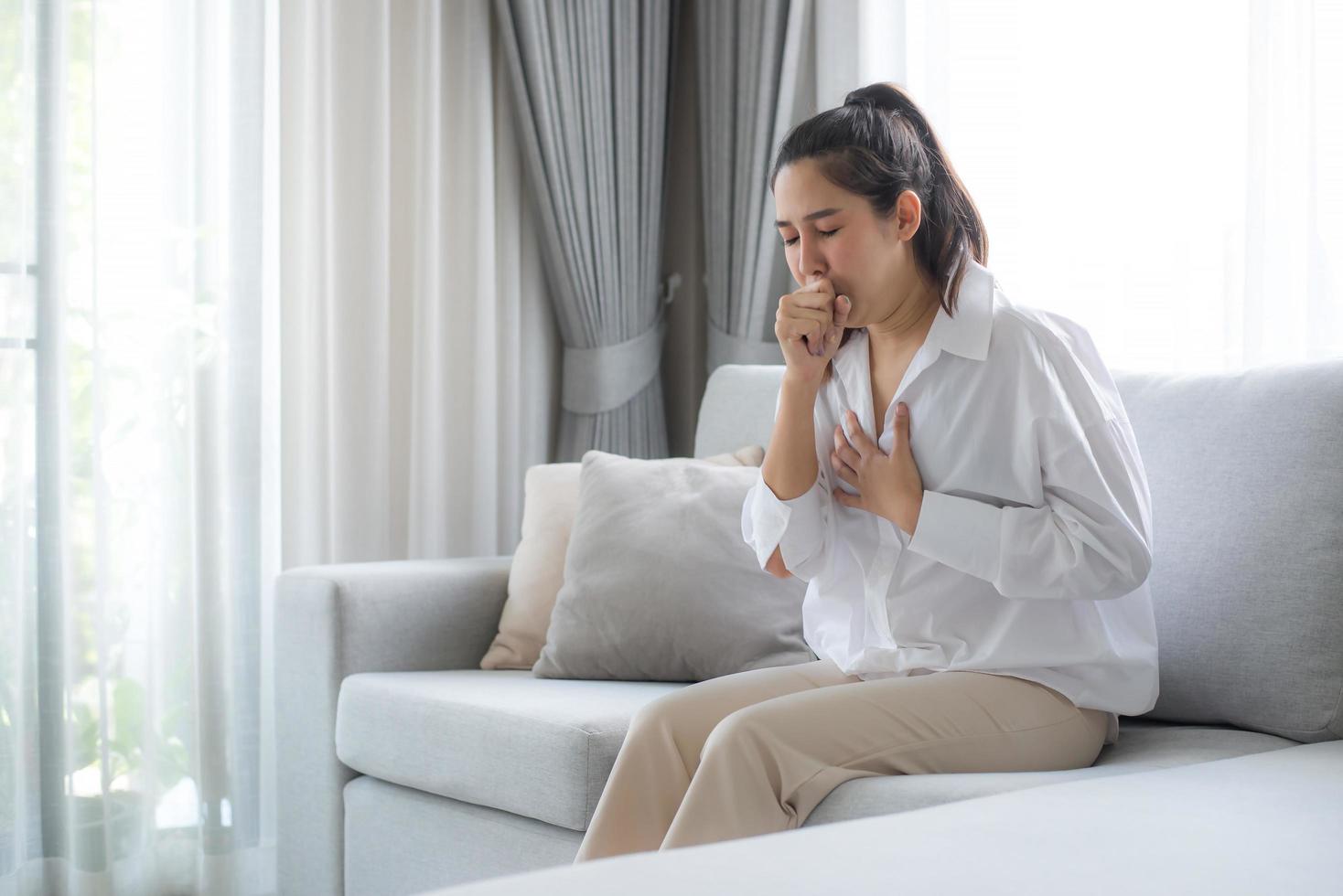 A young woman in a white shirt sits on the sofa in the living room at home. She put her hand on her chest because of a cough, sore throat and discomfort after several days of hard work. photo