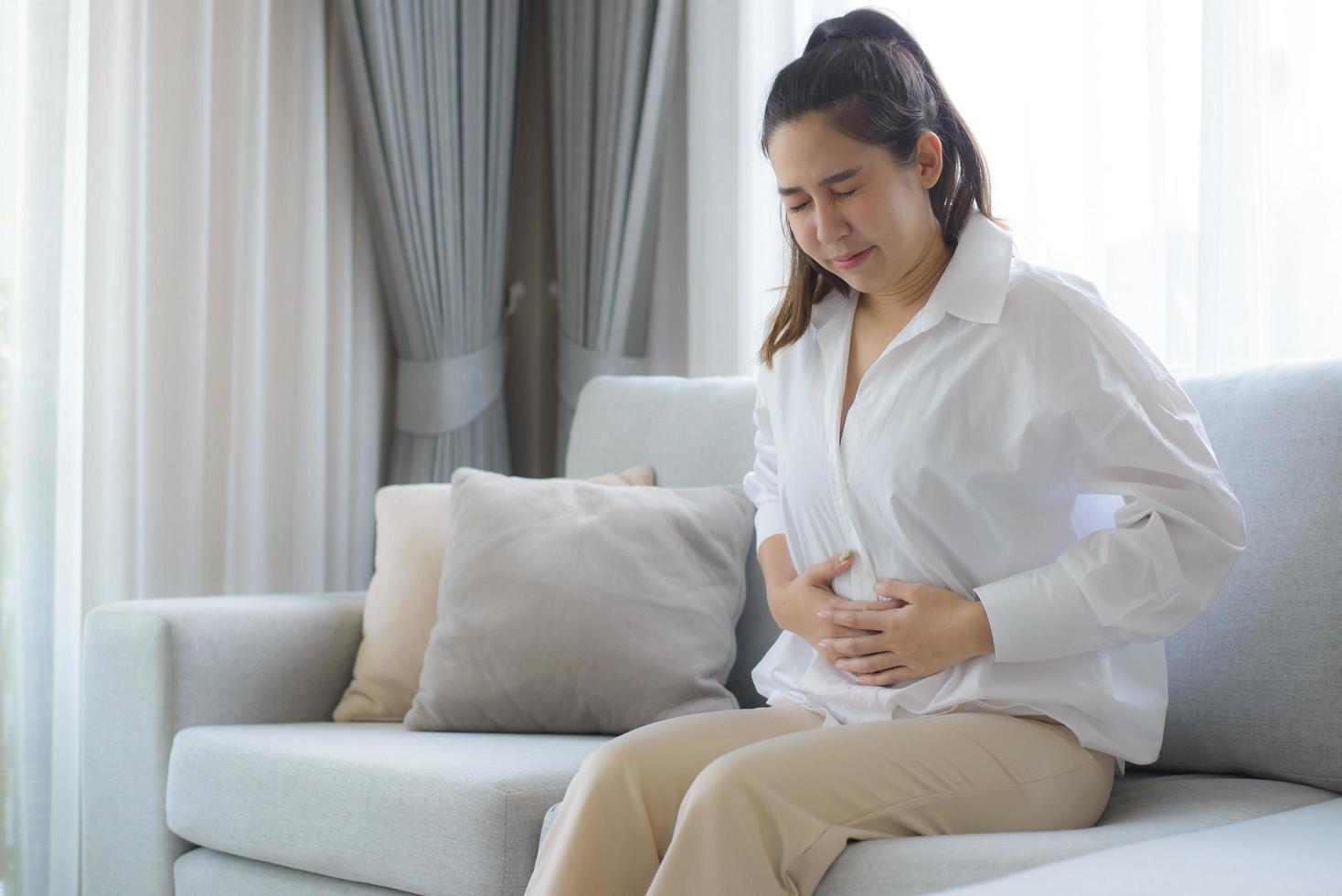 una mujer con un vestido blanco estaba sentada en el sofá de la sala de estar con las manos cruzadas en el estómago. le dolía tanto el estómago que tuvo que ir al hospital. foto